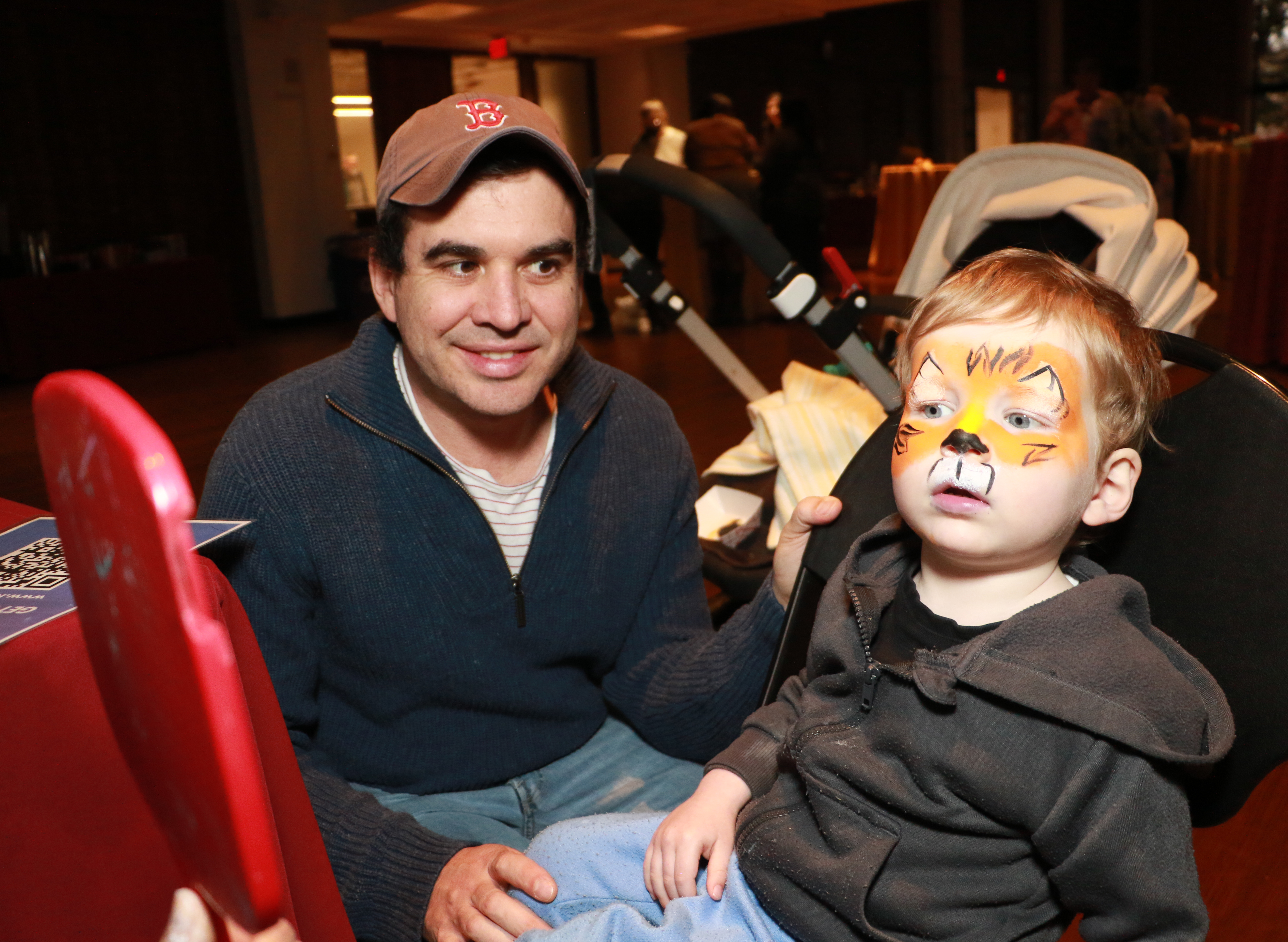 Parent in a baseball cap and grey fleece with a child having their face painted in a stroller