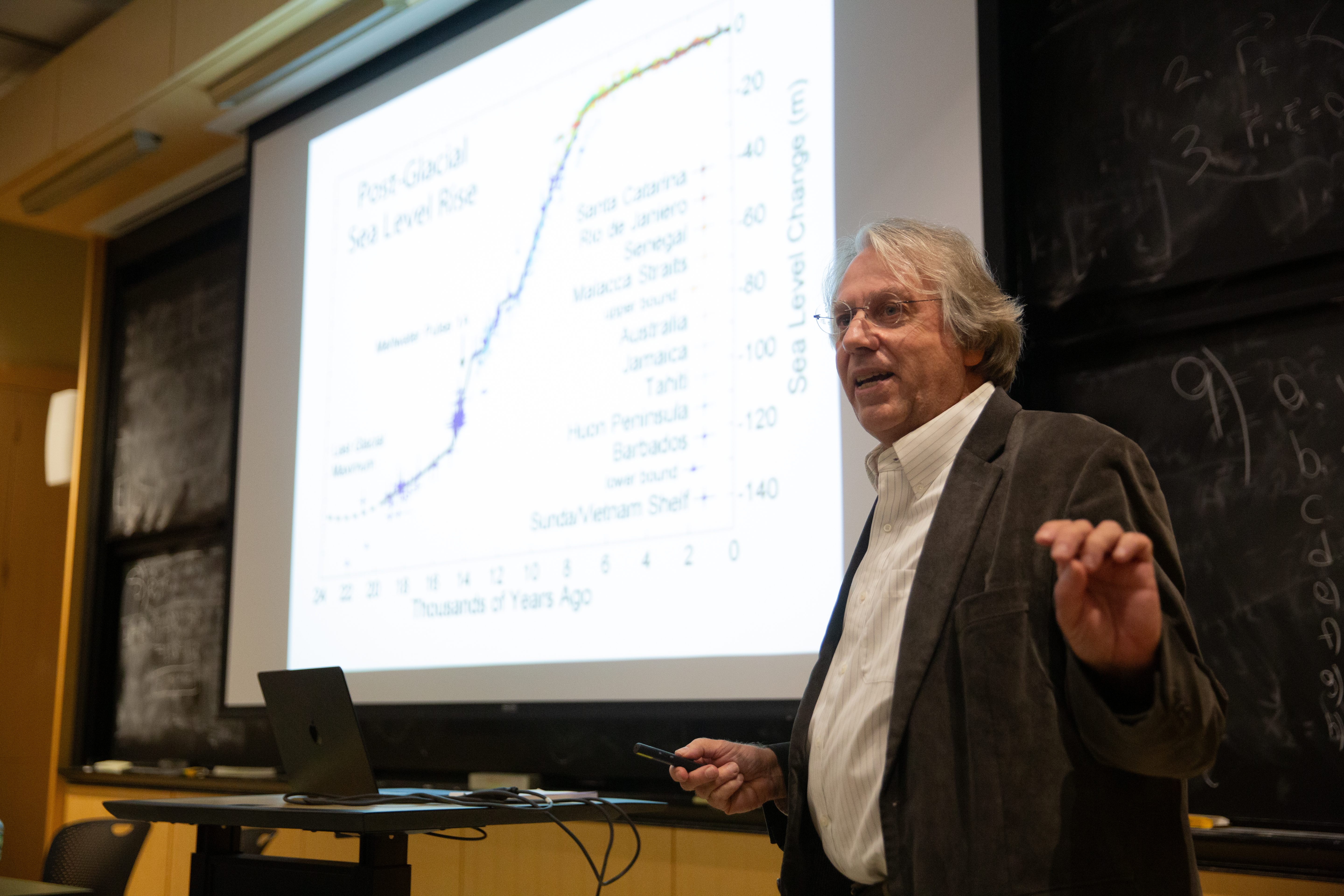Kerry Emanuel discusses sea level rise during a conversation on climate change. The screen behind him features an image plotting the course of sea level rise over millennia.