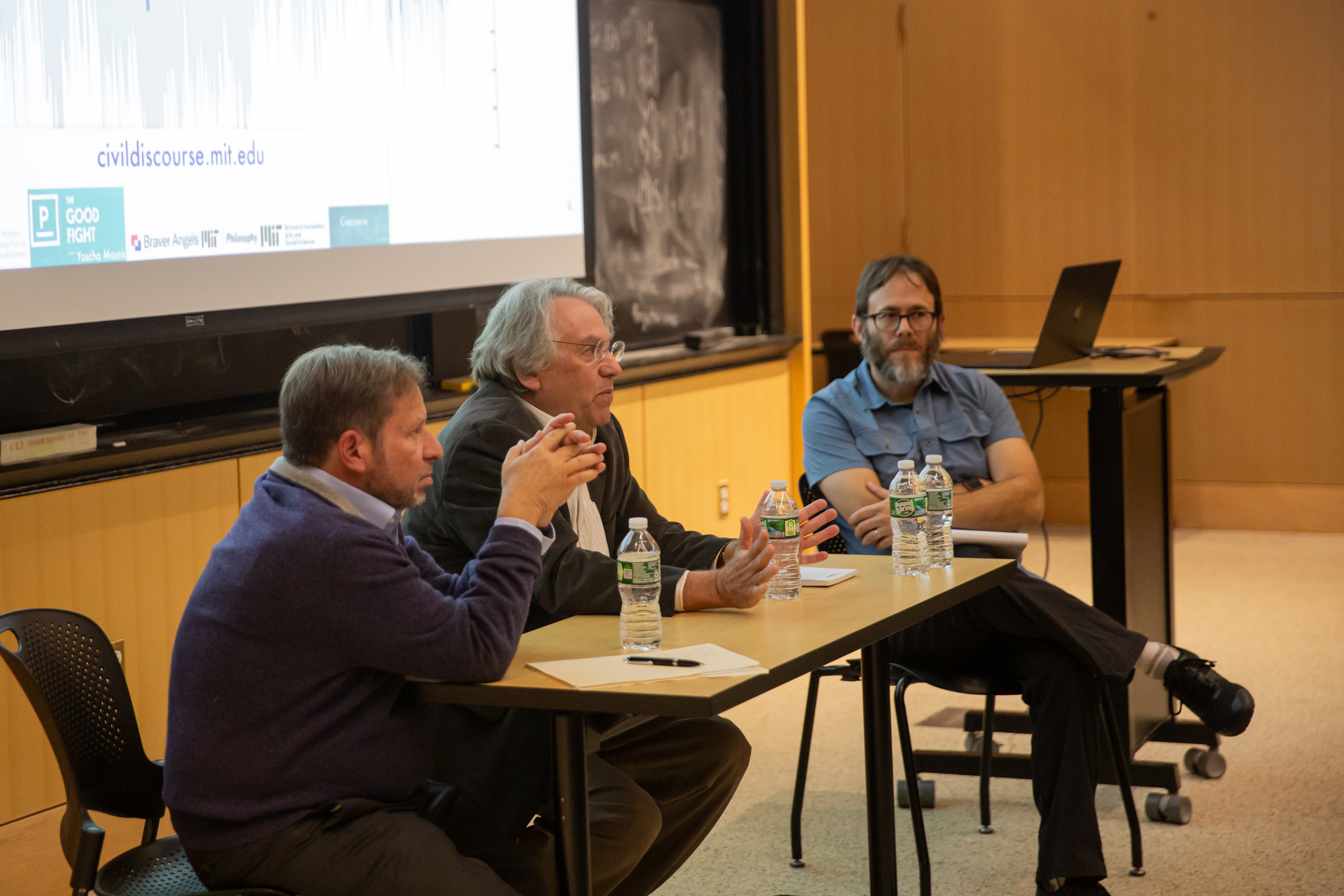 (from left) Steve Koonin, Kerry Emanuel, and moderator Brad Skow discuss climate change with an audience. There's a slide visible on the wall behind them with a web address, civildiscourse.mit.edu, visible. There are bottles of water on the table.