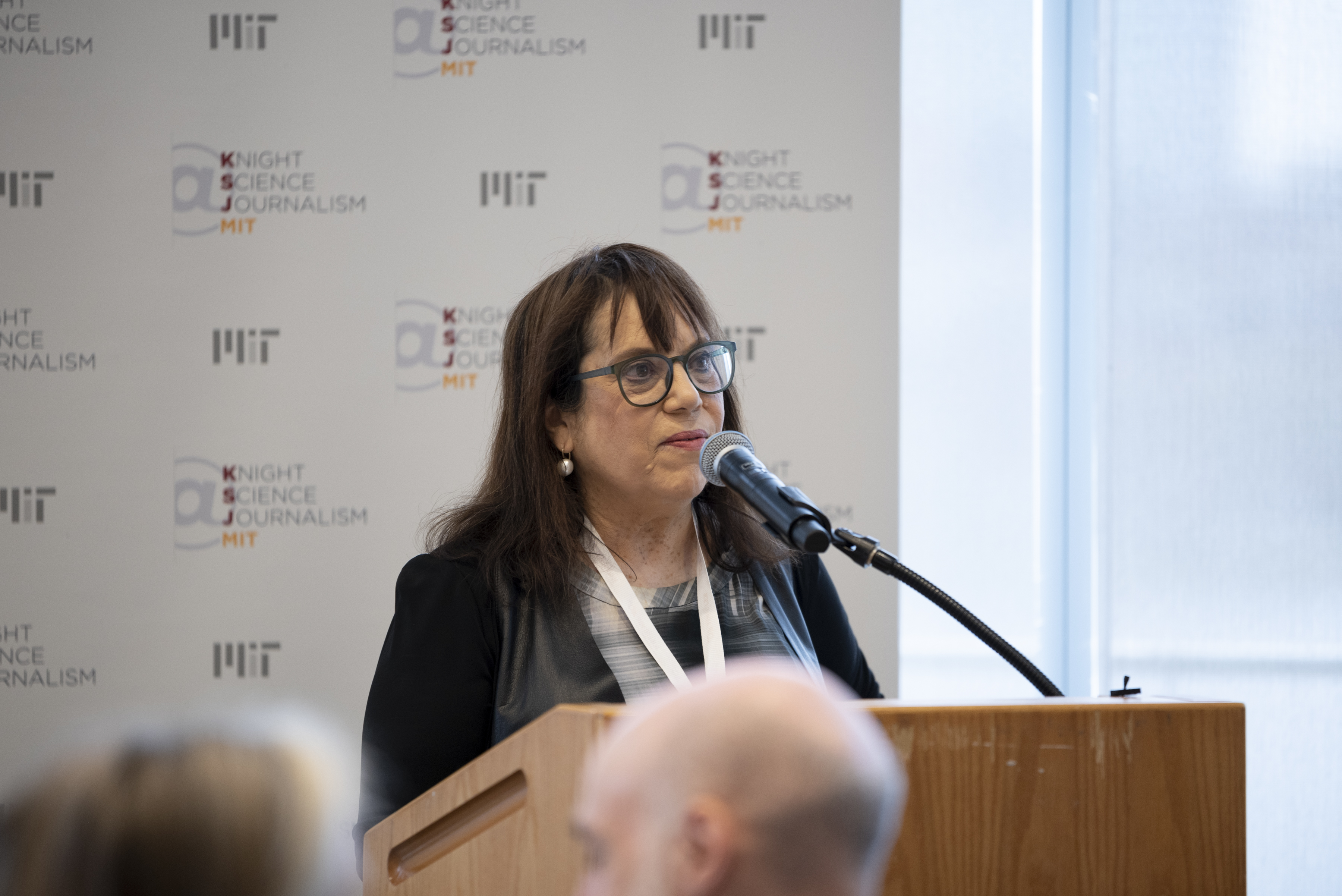 Knight Science Journalism Program Director Deborah Blum addresses the audience at the Victor K. McElheny Awards. She's wearing a black jacket with a satin collar and grey print scoop-necked shirt. She's standing in front of a popup with Knight Science Journalism at MIT branding.