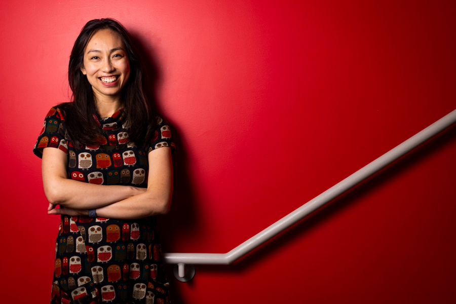 PhD candidate Lisa Ho ’17, who studies barriers that limit women’s participation in the labor force. She's wearing a print dress, smiling, and leaning on a red wall.